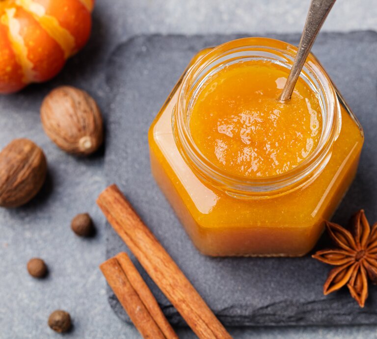 Ein Glas mit Kürbis-Orangen-Marmelade steht auf einer Steinplatte auf einem grauen Untergrund. Neben dem Glas liegt Anis und zwei Zimtstangen. Neben der Steinplatten liegen einige Nüsse und ein Zierkürbis.  | © Getty Images / AnnaPustynnikova