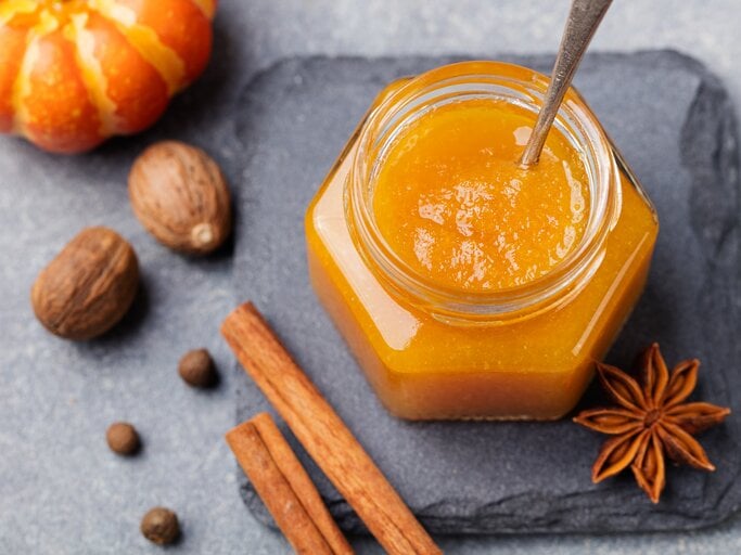 Ein Glas mit Kürbis-Orangen-Marmelade steht auf einer Steinplatte auf einem grauen Untergrund. Neben dem Glas liegt Anis und zwei Zimtstangen. Neben der Steinplatten liegen einige Nüsse und ein Zierkürbis.  | © Getty Images / AnnaPustynnikova