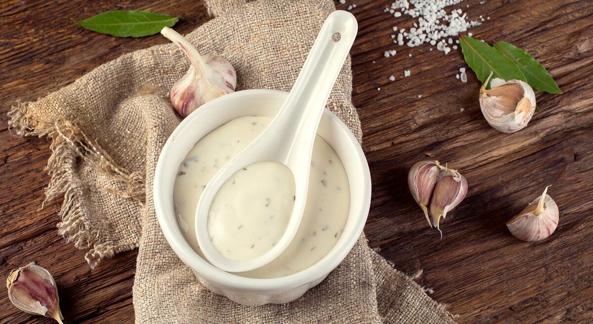 Weiße Schale mit einer cremigen Aioli mit Kräutern auf einem Holzbrett.
