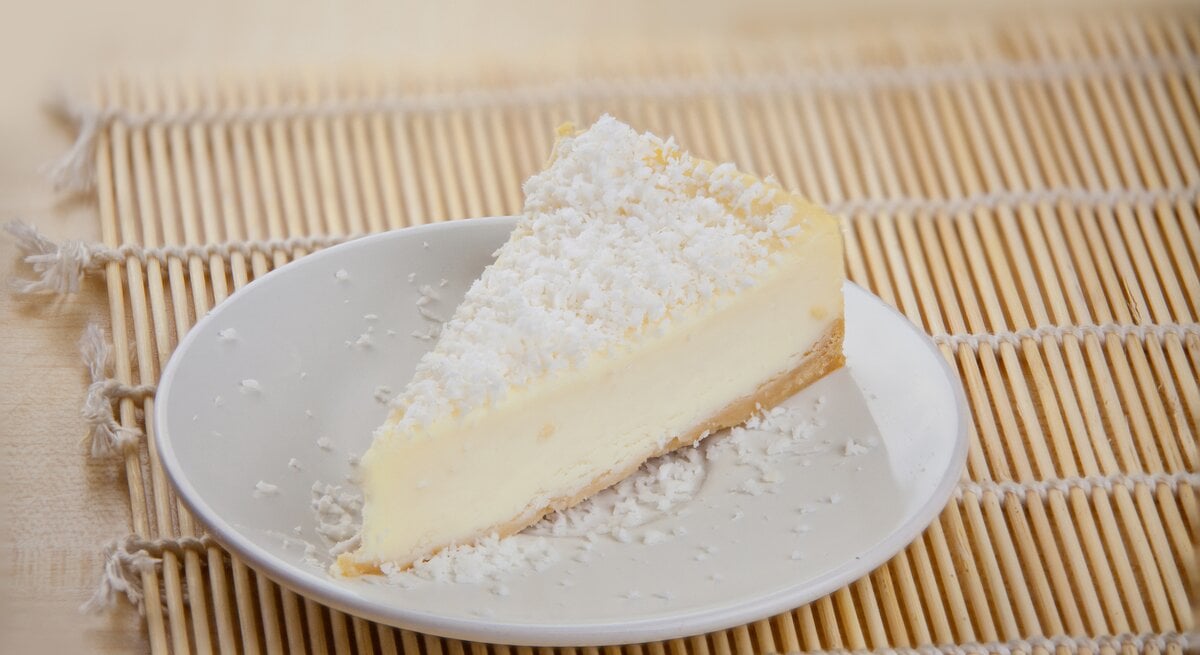 Ein Stück cremiger Joghurtkuchen mit Kokosraspeln bestreut auf einem weißen Teller, der auf einer Bambusmatte steht.