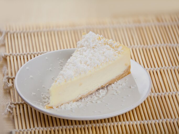 Ein Stück cremiger Joghurtkuchen mit Kokosraspeln bestreut auf einem weißen Teller, der auf einer Bambusmatte steht. | © iStock/Nikolaeva Galina