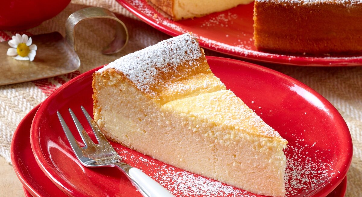 Ein Stück Käsekuchen auf einem roten Teller und hellem Holztisch. Alles von schräg oben fotografiert.
