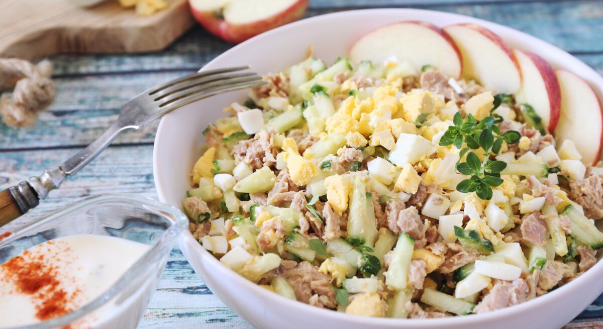 Cremiger Salat aus Ei, Thunfisch und Gurken, mit Apfelscheiben und Kräutern garniert, serviert auf einem Holztisch.