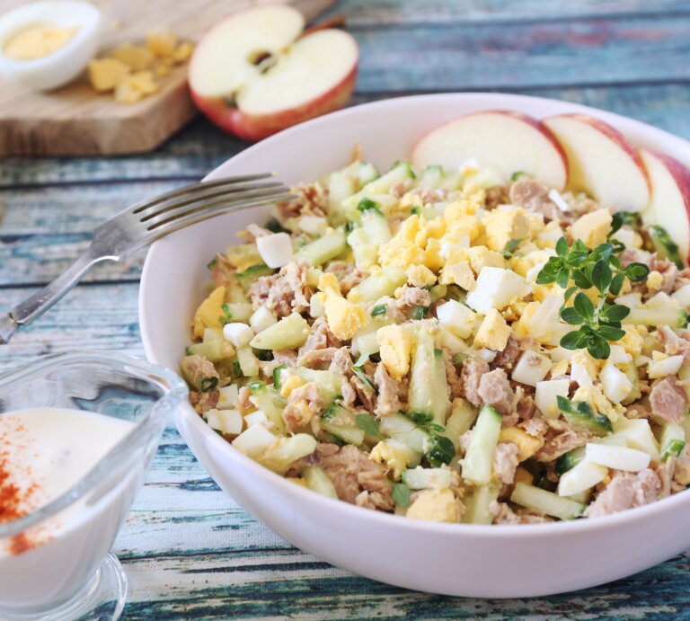 Cremiger Salat aus Ei, Thunfisch und Gurken, mit Apfelscheiben und Kräutern garniert, serviert auf einem Holztisch. | © iStock.com/photosimysia