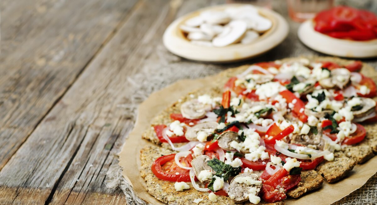 Die Chiasamen-Pizza auf einem rustikalem Holztisch von schräg oben fotografiert. Im Hintergrund noch unscharf Zutaten zum Belegen zu erkennen.