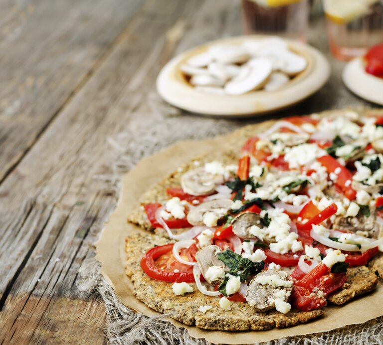 Die Chiasamen-Pizza auf einem rustikalem Holztisch von schräg oben fotografiert. Im Hintergrund noch unscharf Zutaten zum Belegen zu erkennen. | © iStock/nata_vkusidey