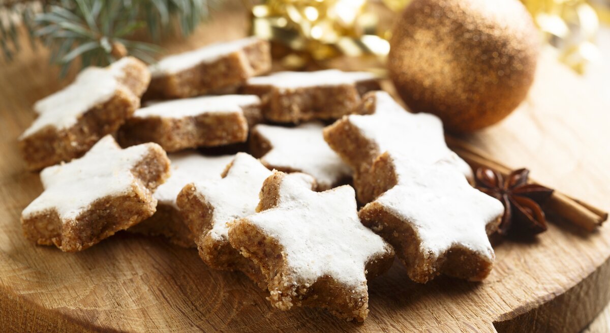 Mehrere frisch gebackene Zimtsterne auf einem Holzbrett. Daneben weihnachtliche Dekorationselemente wie Tannenzweige und goldenes Lametta. 