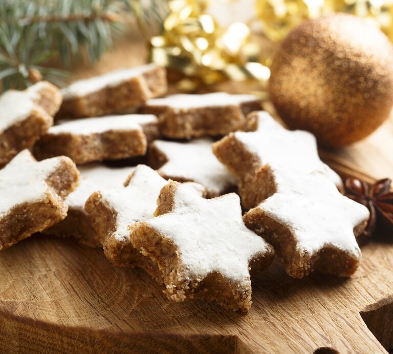 Mehrere frisch gebackene Zimtsterne auf einem Holzbrett. Daneben weihnachtliche Dekorationselemente wie Tannenzweige und goldenes Lametta.  | © iStock/ Mariha-kitchen