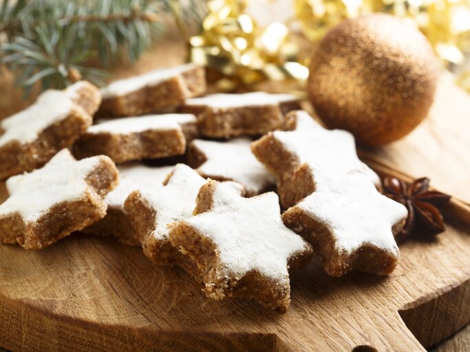 Mehrere frisch gebackene Zimtsterne auf einem Holzbrett. Daneben weihnachtliche Dekorationselemente wie Tannenzweige und goldenes Lametta.  | © iStock/ Mariha-kitchen