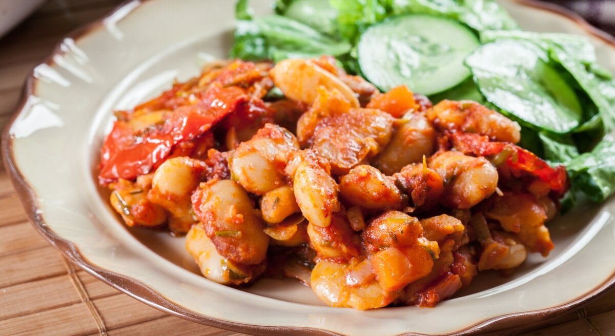 Weiße Bohnen in Tomatensoße mit Gurkensalat auf weißem Teller. 