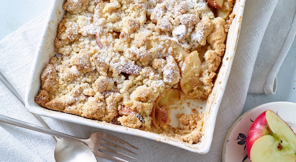 Apple Crumble auf weißem Tuch, daneben Äpfel und Besteck.