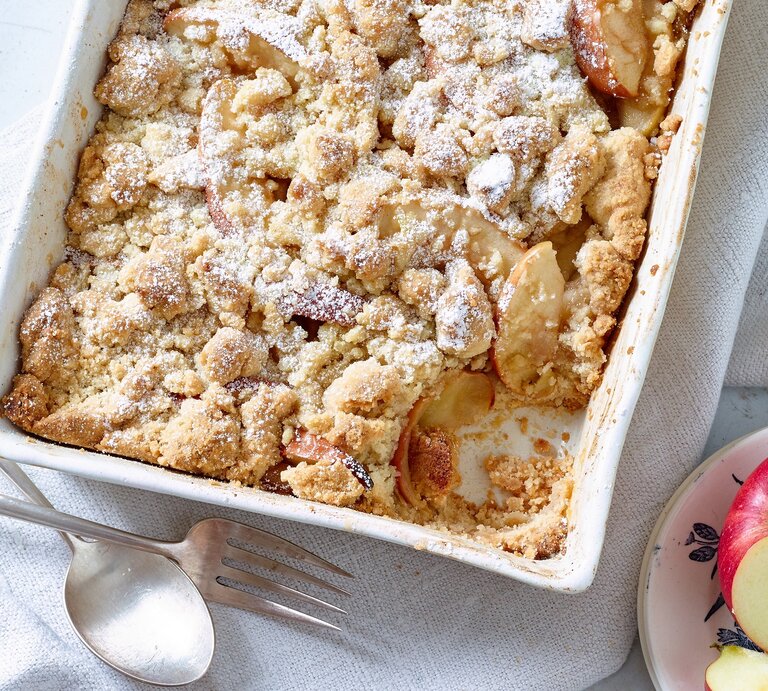 Apple Crumble auf weißem Tuch, daneben Äpfel und Besteck. | © Matthias Liebich