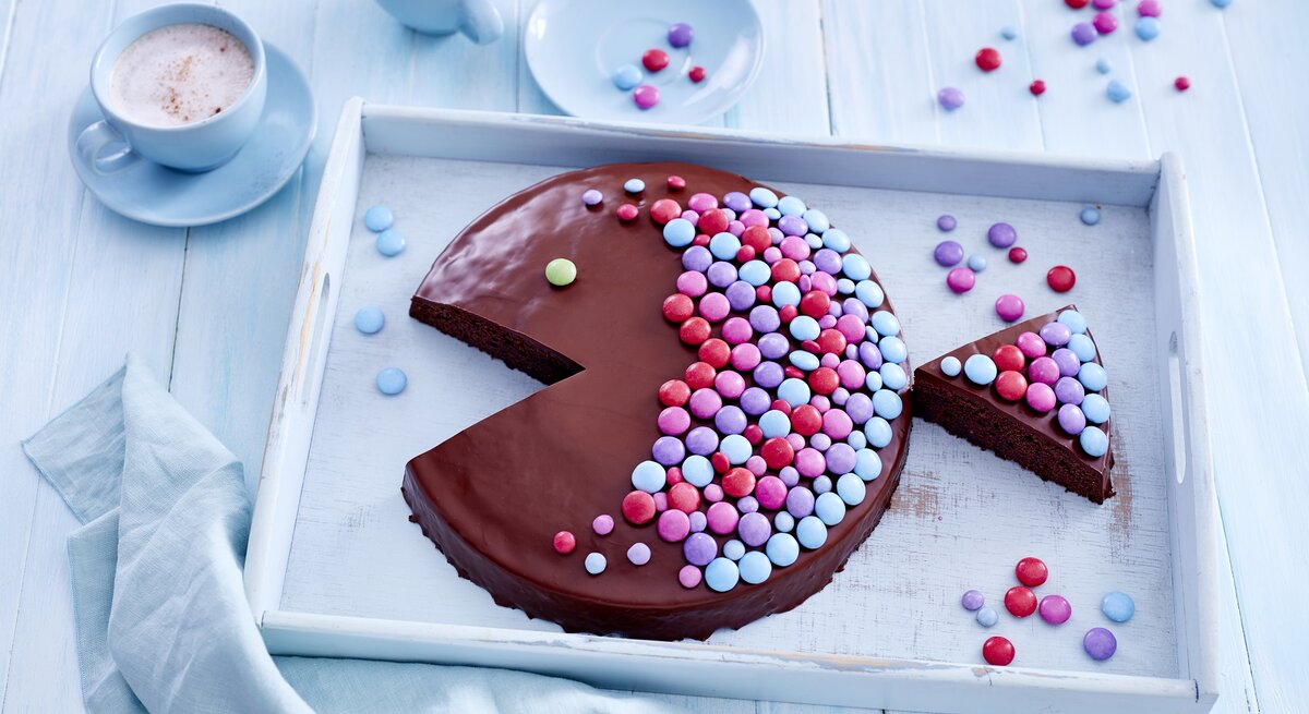 Ein Schokoladen-Kuchen in Fischform der mit bunten Streuseln beleget ist auf einem hellblauen Untergrund und weißem Tablett von oben fotografiert.
