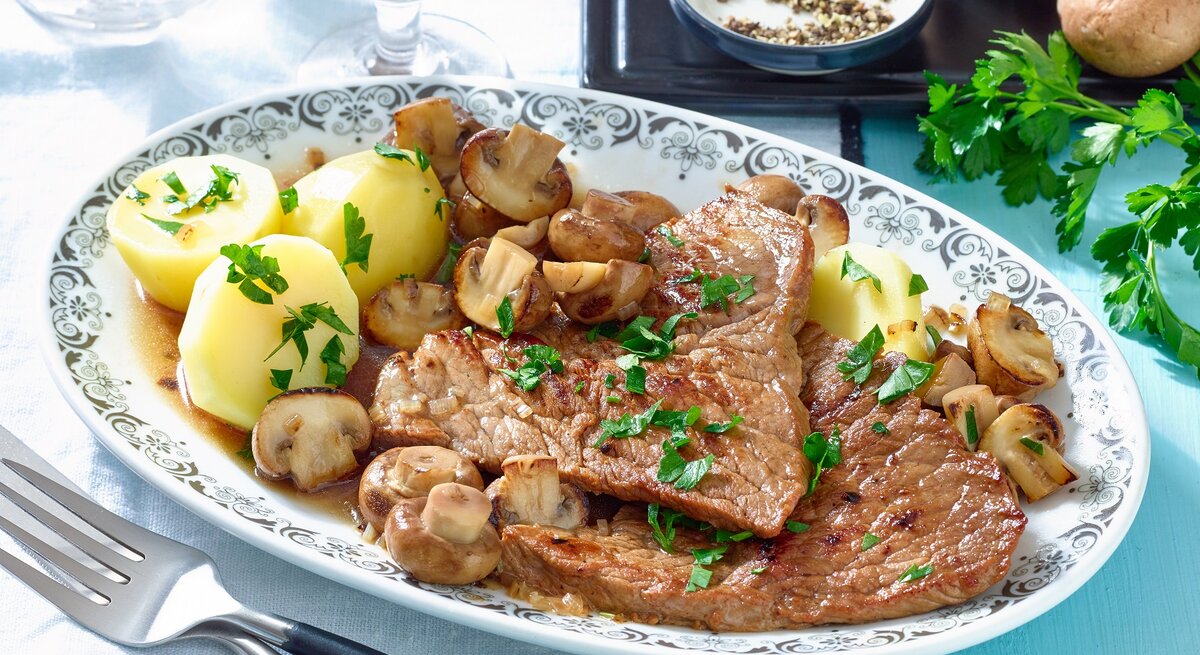 Schnitzel mit Champignons und Kartoffeln / ovaler Teller aus Porzellen/ Gabel / frische Petersilie / kleine runde Schale mit Pfefferkörnern / zwei Gläser / zwei frische Champignons / Draufsicht, schräg