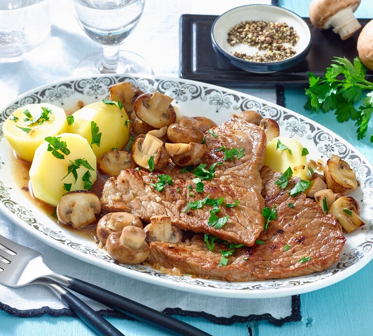 Schnitzel mit Champignons und Kartoffeln / ovaler Teller aus Porzellen/ Gabel / frische Petersilie / kleine runde Schale mit Pfefferkörnern / zwei Gläser / zwei frische Champignons / Draufsicht, schräg | © Matthias Liebich