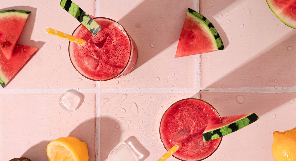 Wassermelone Frosé in zwei großen Gläsern mit Strohhalm und frischer Melone auf einem rosa Tisch aus Fliesen. 