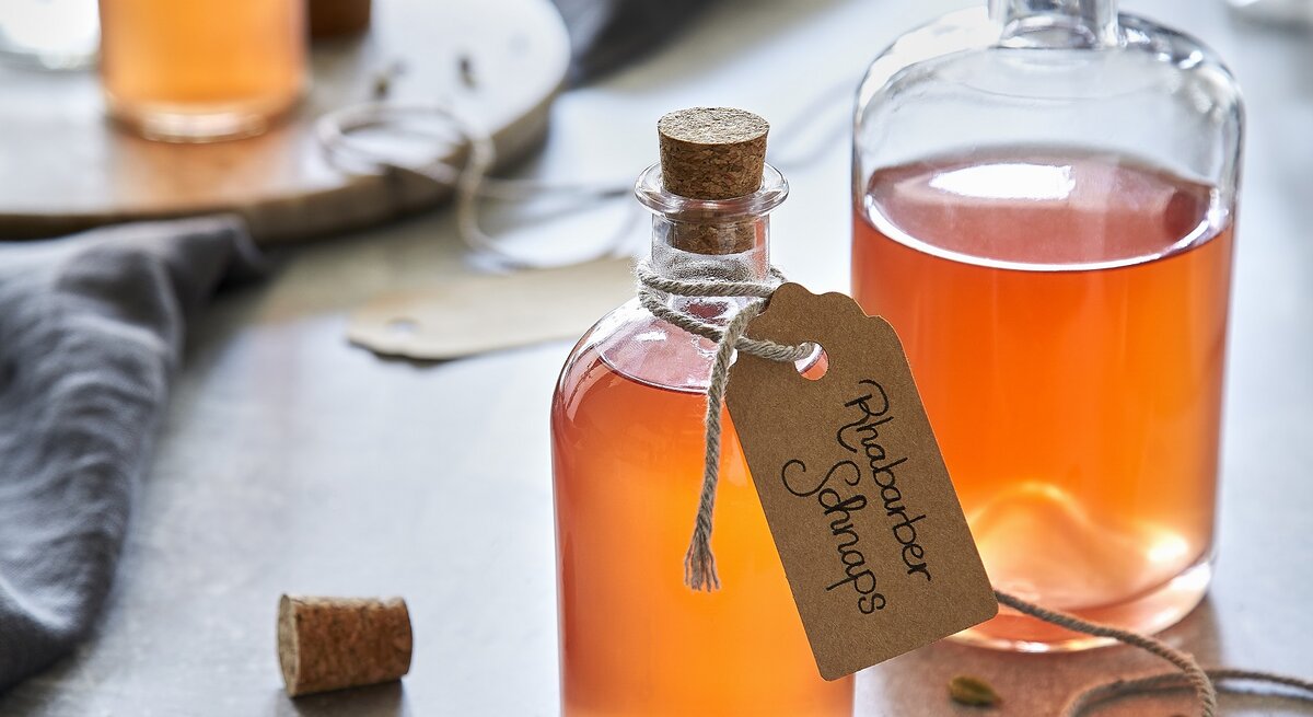Draufsicht schräg: Drei Flaschen aus Glas in denen eine Flüssigkeit ist. Um eine Flasche hängt ein kleines Papierschild, auf dem "Rhabarberschnaps" geschrieben steht.