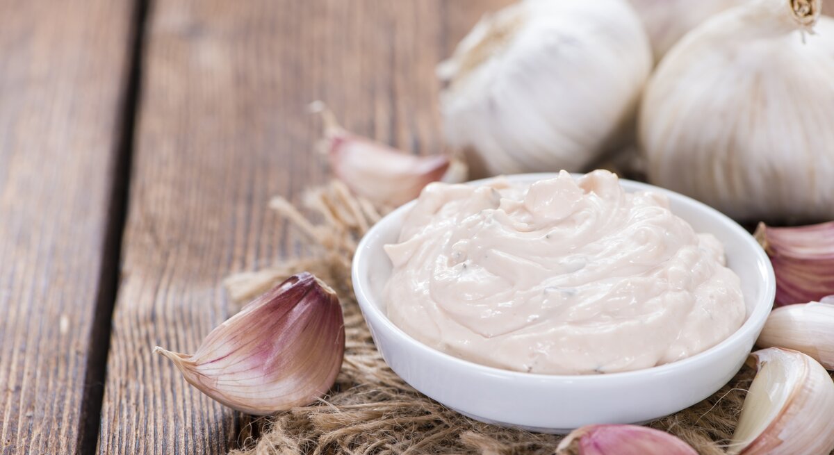 Schale mit Aioli auf Holztisch, daneben Knoblauchzehen. 
