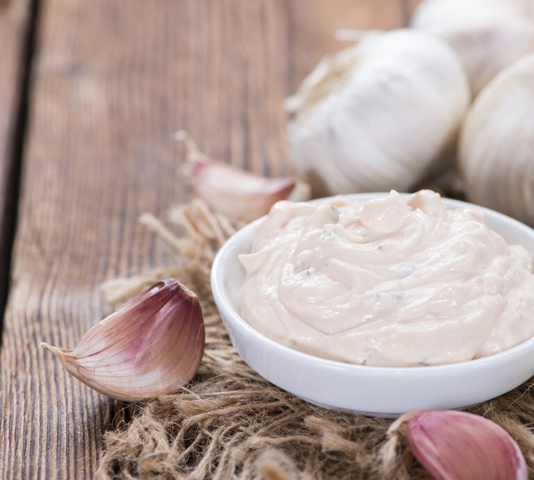 Schale mit Aioli auf Holztisch, daneben Knoblauchzehen.  | © iStock/ HandmadePictures