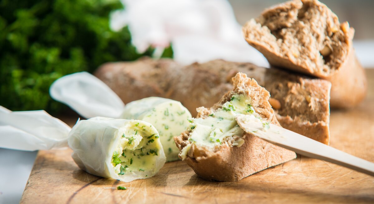 Hausgemachte Kräuterbutter eingewickelt in Brotpapier, angeschnitten auf einem Holzbrett. Daneben mehrere Scheiben Weißbrot.