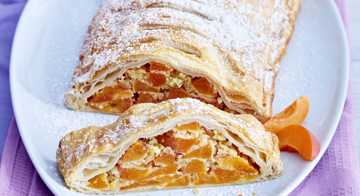 Aprikosen-Mandel-Strudel mit Puderzucker bestreut und angeschnitten serviert auf einem weißen Teller auf rosanem Untergrund. 