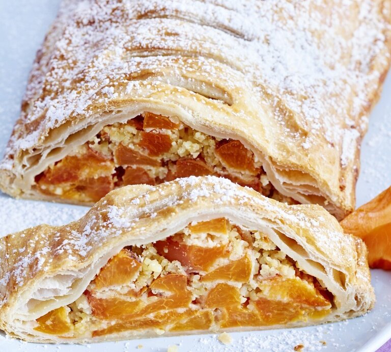Aprikosen-Mandel-Strudel mit Puderzucker bestreut und angeschnitten serviert auf einem weißen Teller auf rosanem Untergrund.  | © EAT CLUB /Isabell Triemer