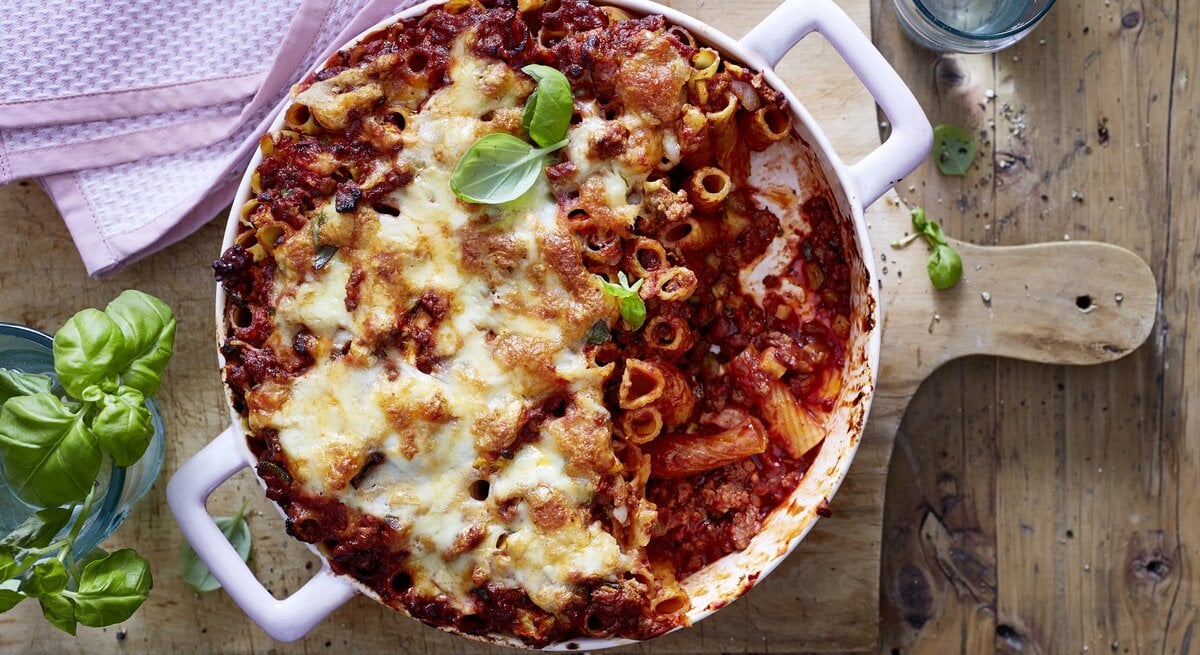 Rigatoni-Auflauf mit Hacksoße in einer weißen Auflaufform auf einem Holzbrett.