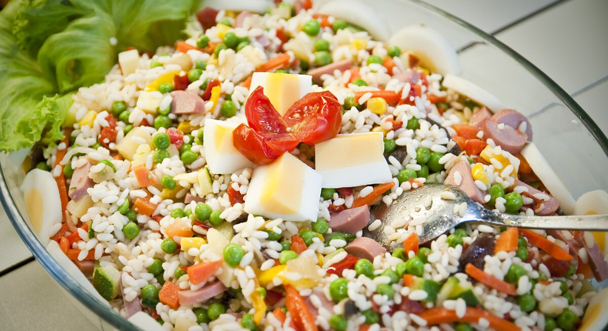 Kunterbunter Reissalat mit Paprika, Würstchen, gekochtem Ei und Erbsen in Glasschüssel. 