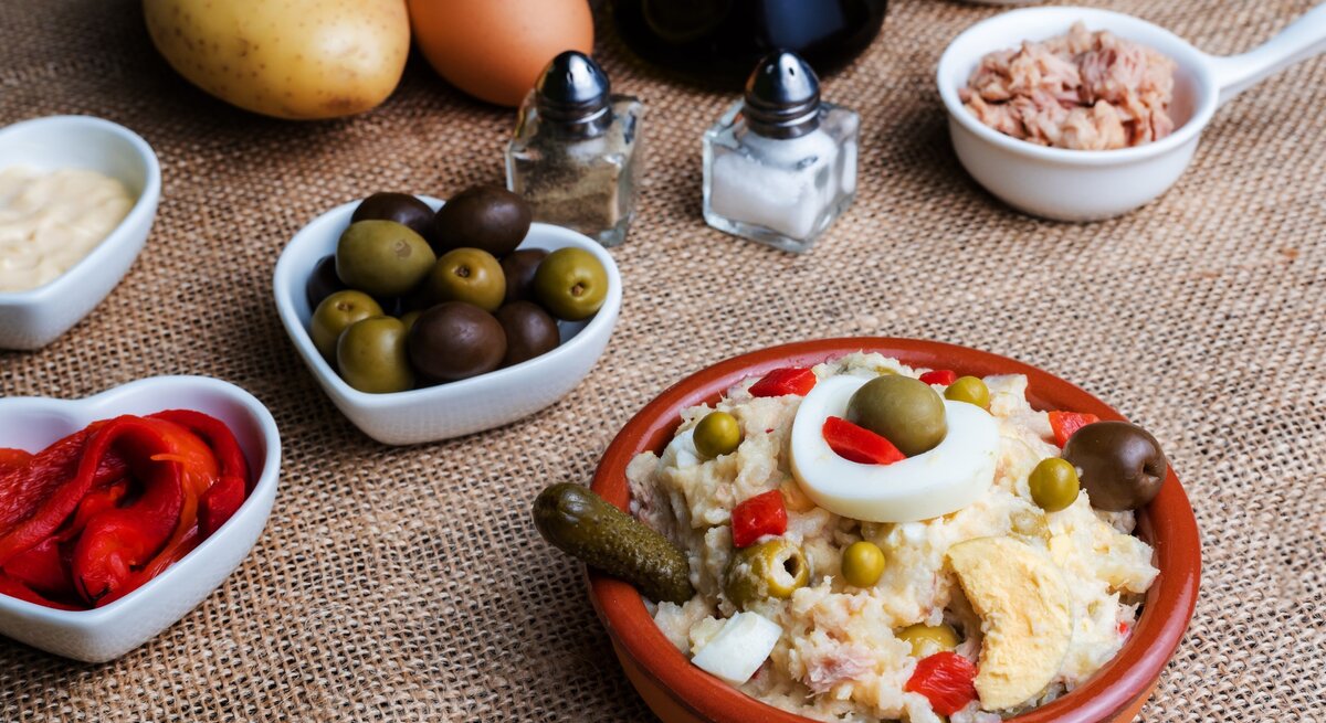 Spanischer Kartoffelsalat Ensalada Rusa in Tapasschale mit Tapas und Zutaten auf hellbrauner Tischdecke. Draufsicht.