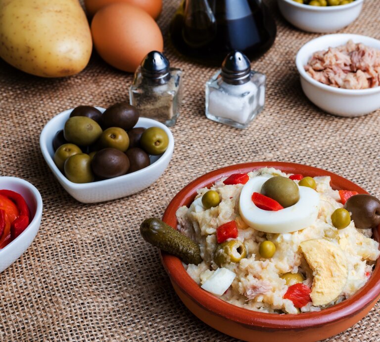 Spanischer Kartoffelsalat Ensalada Rusa in Tapasschale mit Tapas und Zutaten auf hellbrauner Tischdecke. Draufsicht. | © Getty Images / Miguel Perfectti
