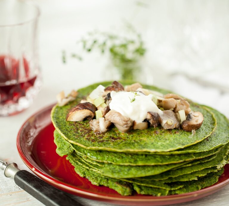 Bärlauch-Pfannkuchen mit Champignons und Crème Fraiche auf einem roten Teller mit einem Glas Rotwein auf einem weißen Holztisch.  | © GettyImages/ Carola Fink