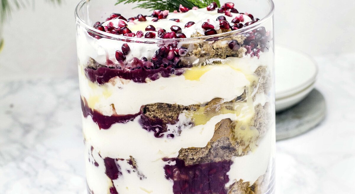 Das Lebkuchen-Trifle mit Eierlikör im Glas vor hellem Untergund mit weihnachtlicher Deko garniert. Alles sehr nah und direkt von vorne fotografiert.