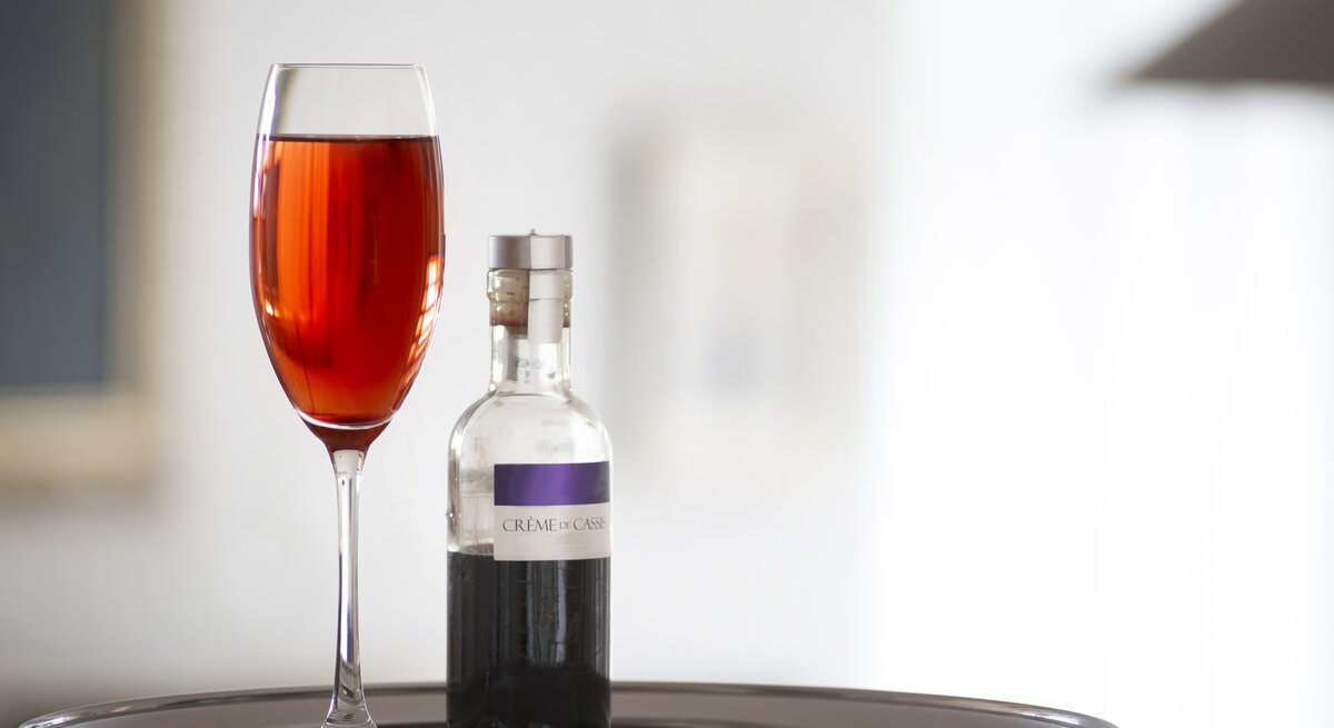Ein Glas mit dem Kir Royal vor hellem Hintergrund und daneben eine Flasche mit dem Cassis-Sirup.