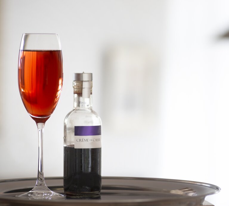 Ein Glas mit dem Kir Royal vor hellem Hintergrund und daneben eine Flasche mit dem Cassis-Sirup. | © iStock/ AM-C
