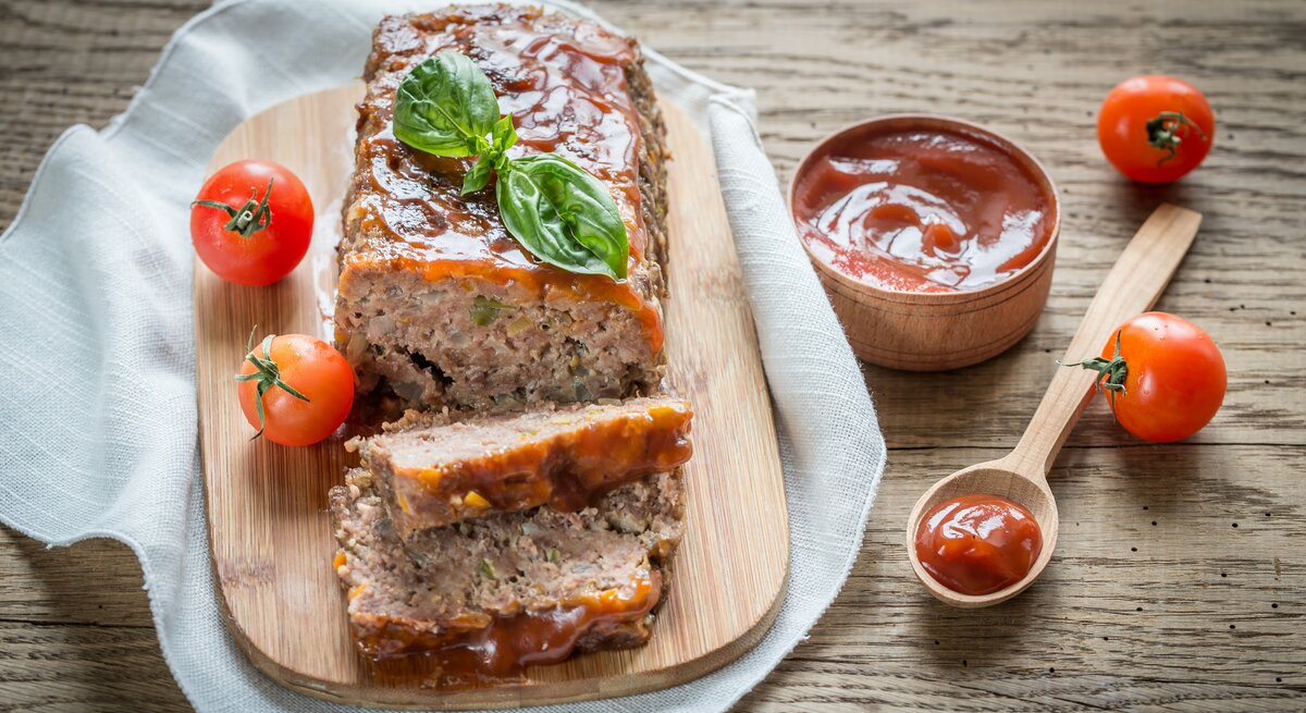 Ein Lieblingshackbraten mit Tomaten-Rotwein-Überzug liegt auf einem Holzbrett auf einem Geschirrtuch. Um ihn  herum liegen ein Löffel, Tomaten und eine Schale mit Tomaten-Rotwein-Mischung.