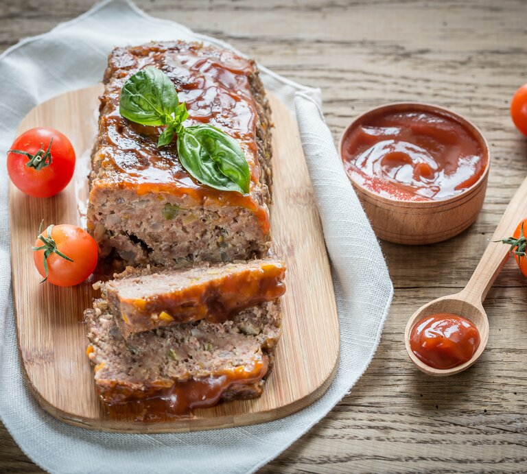 Ein Lieblingshackbraten mit Tomaten-Rotwein-Überzug liegt auf einem Holzbrett auf einem Geschirrtuch. Um ihn  herum liegen ein Löffel, Tomaten und eine Schale mit Tomaten-Rotwein-Mischung. | © Shutterstock / Alexander Prokopenko