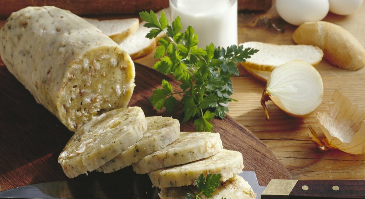 Auf einem rustikalen Holzbrett liegen aufgeschnittene Böhmische Knödel, dekoriert mit frischer Petersilie. Daneben stehen Brot, Eier und eine halbierte Zwiebel.