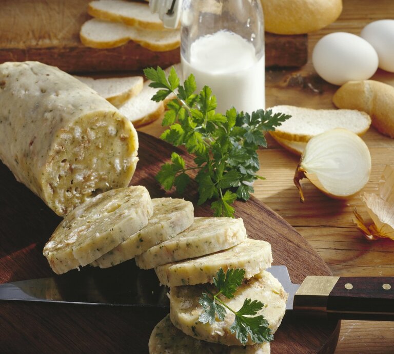 Auf einem rustikalen Holzbrett liegen aufgeschnittene Böhmische Knödel, dekoriert mit frischer Petersilie. Daneben stehen Brot, Eier und eine halbierte Zwiebel. | © StockFood/Teubner Foodfoto GmbH