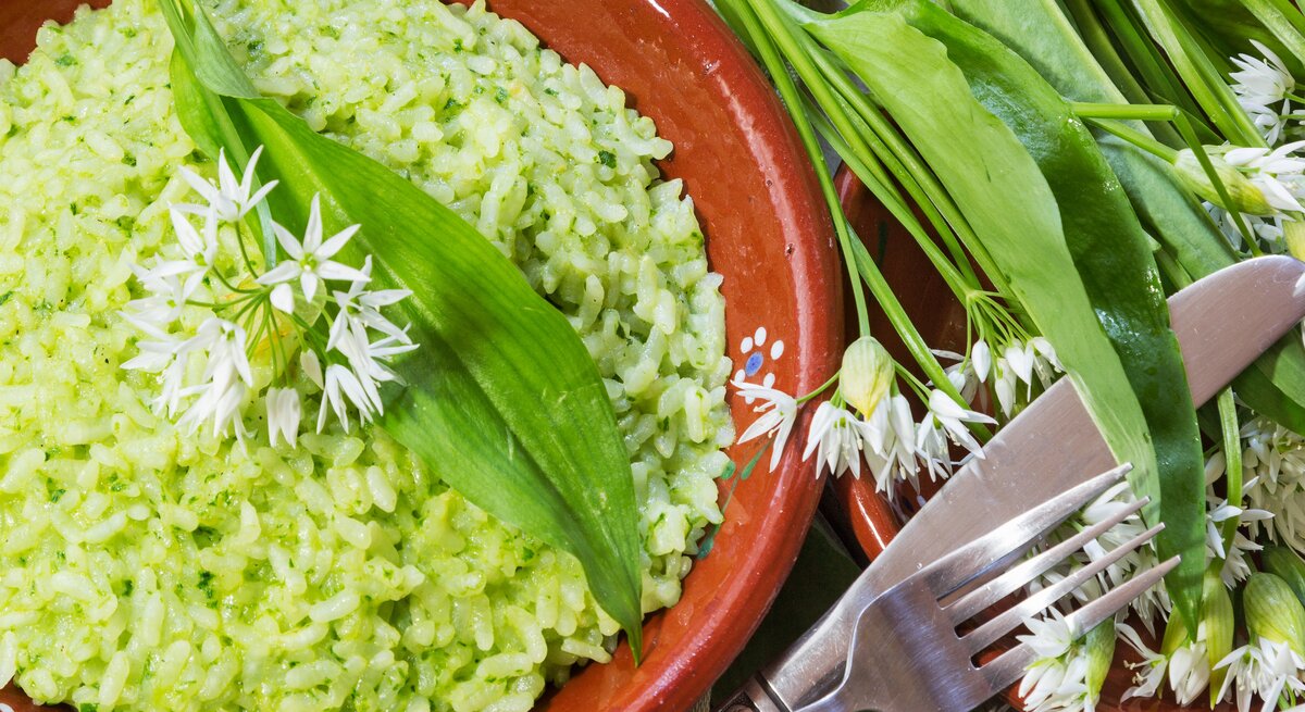 Grünes Bärlauch-Risotto in einem rötlichen Teller aus Ton neben frischen Bärlauch-Blättern auf einem hellen Holztisch mit Besteck von oben.