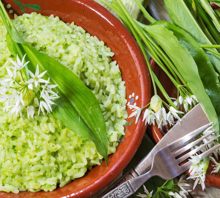 Grünes Bärlauch-Risotto in einem rötlichen Teller aus Ton neben frischen Bärlauch-Blättern auf einem hellen Holztisch mit Besteck von oben. | © GettyImages/ Yu Lan