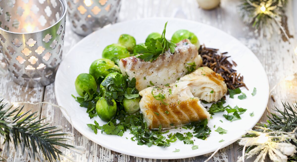 Ein festlich dekoriertes Tellergericht mit gebratenem Fisch, frischen Rosenkohl und Wildreis, garniert mit frischen Kräutern, auf einem rustikalen Holztisch. Im Hintergrund sind weihnachtliche Dekorationen wie Zweige und Kerzen zu sehen.