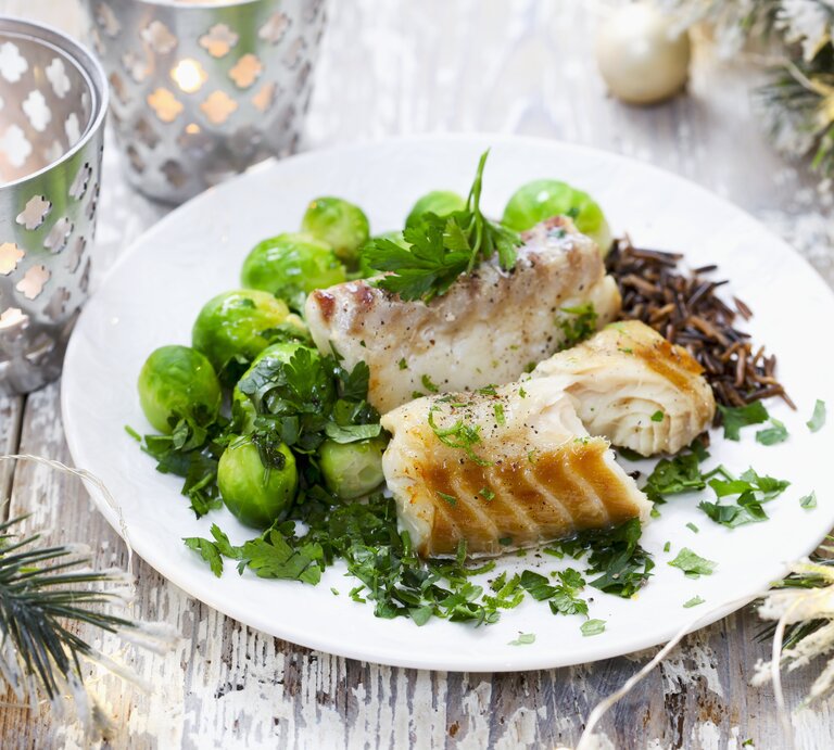 Ein festlich dekoriertes Tellergericht mit gebratenem Fisch, frischen Rosenkohl und Wildreis, garniert mit frischen Kräutern, auf einem rustikalen Holztisch. Im Hintergrund sind weihnachtliche Dekorationen wie Zweige und Kerzen zu sehen. | © StockFood / Bialy, Boguslaw