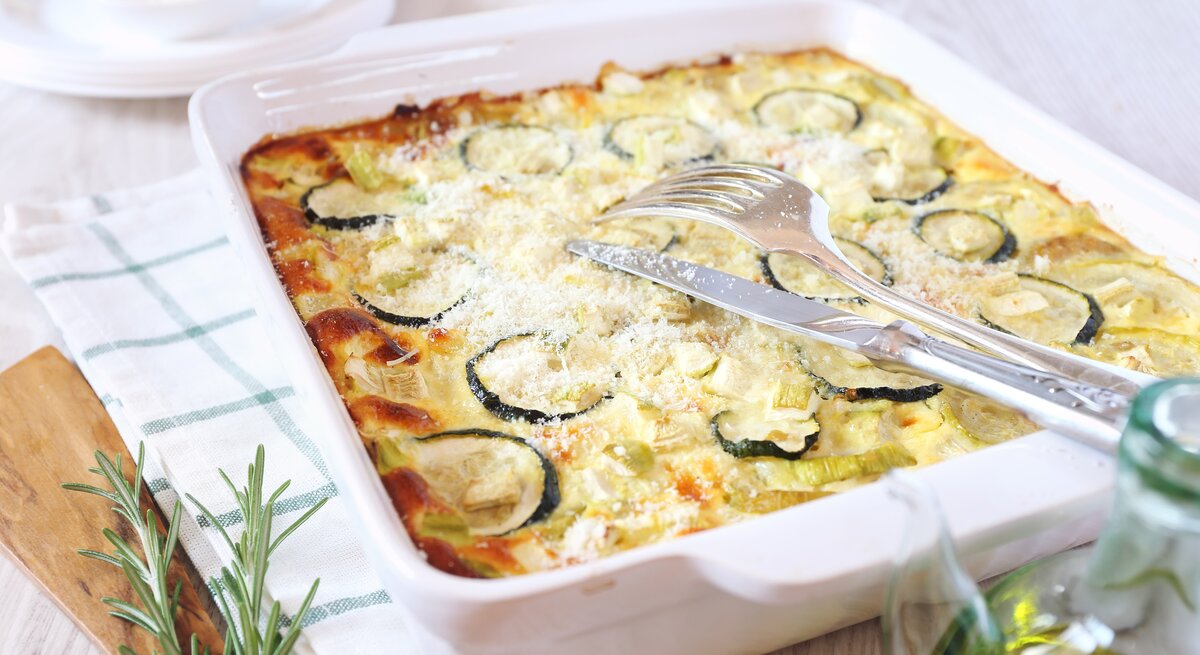 Auflaufform gefüllt mit Zucchini-Auflauf angerichtet auf mediterranem Geschirrtuch. 