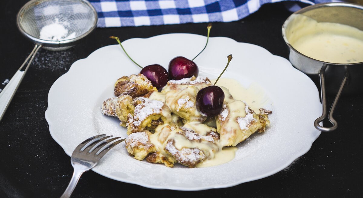 Kaiserschmarrn mit Kirschen und Vanillesauce aus einem runden Teller / Sieb mit Puderzucker / Draufsicht, schräg