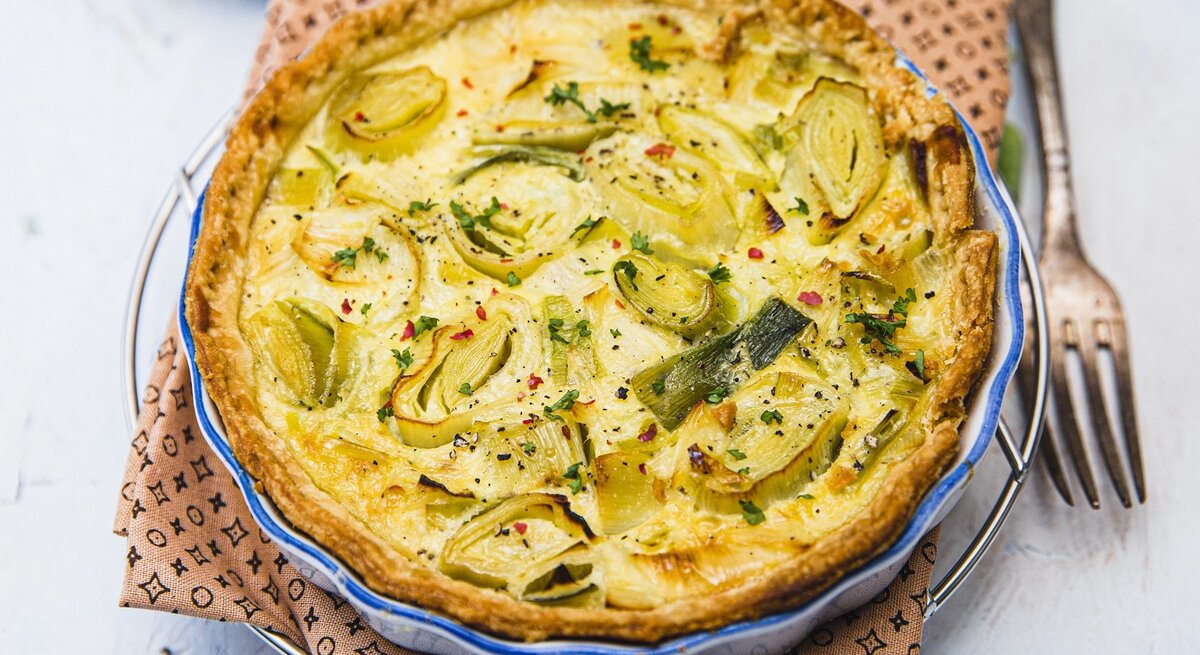 Eine goldbraun gebackene Quiche mit Lauchscheiben in einer runden Backform mit blauem Rand.