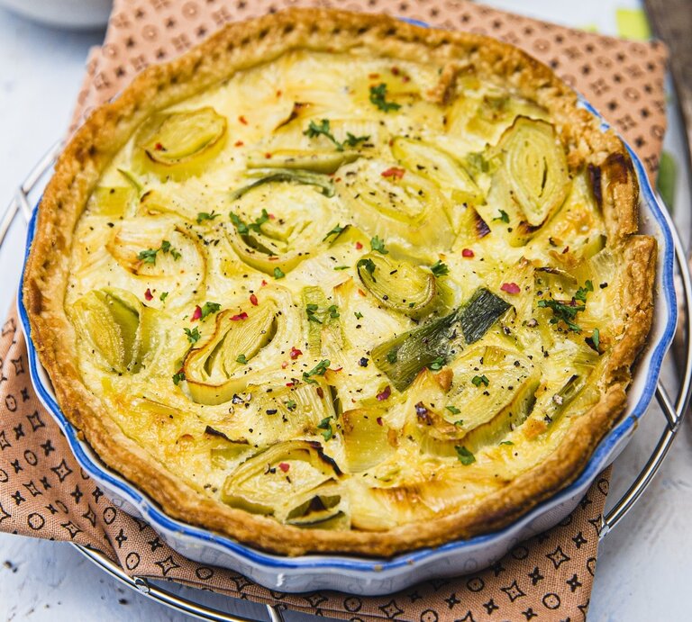 Eine goldbraun gebackene Quiche mit Lauchscheiben in einer runden Backform mit blauem Rand. | © StockFood / PhotoCuisine