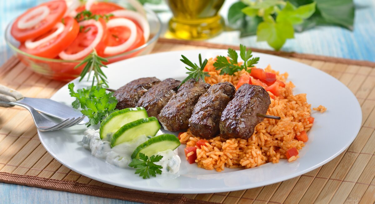 Cevapcici auf Tomatenreis mit Gurkenscheiben und Kräutern auf weißem Teller, daneben ein Tomatensalat und Olivenölflasche.