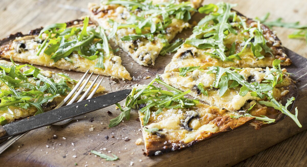 Nahaufnahme vom in Stücke geschnittenen, veganen Flammkuchen mit Rucola und Pilzen.
