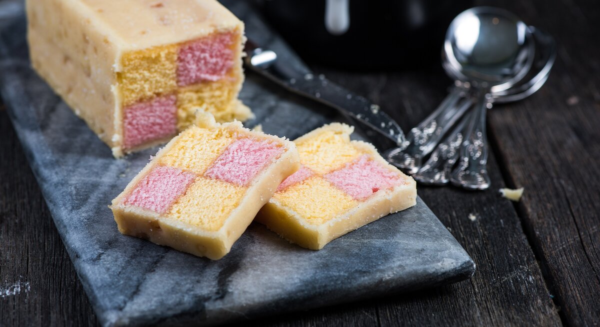 Angeschnittener britischer Battenburg-Kuchen auf einer dunklen Steinplatte auf einem dunklen Holztisch.