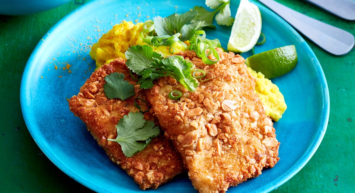 Erdnuss-Schnitzel mit Curry-Stampf auf einem Teller serviert. Rechts davon Besteck. Draufsicht.